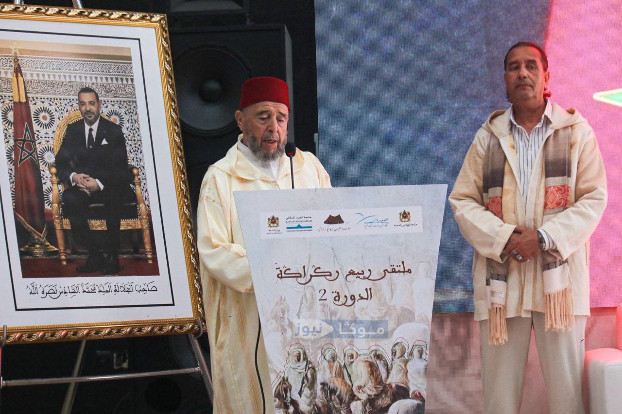 Moroccan man in a red fez speaks at a podium during a cultural event, with a framed portrait of a king on the left and banners nearby.