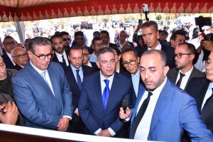 Group of men in suits at an outdoor event, the man in front holding a microphone as others listen and crowd behind them records with phones