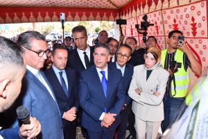 A man in a blue suit speaks to a crowd of officials and press inside a red-and-white patterned tent, with photographers and a woman in a beige suit watching nearby.