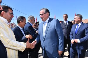 Officials in suits shake hands at an outdoor formal event under a clear blue sky, with others nearby in line and red flags in the background.