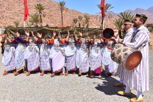 Group of dancers in white lace outfits performing a traditional Moroccan dance outdoors, with two drummers on the right and desert landscape in the background.