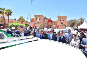 Officials in suits and a crowd gather around a white vehicle during a sunny outdoor event at a tourist information site, as a man in a dark suit points ahead.