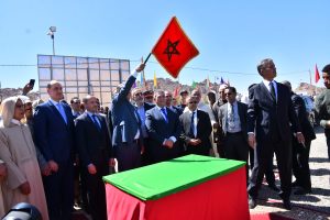 Group of formally dressed people at an outdoor ceremony around a coffin draped in green and red fabric; a man raises a red flag with a black pentagram symbol.