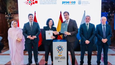 Six officials in suits stand during a signing ceremony; the center pair shake hands and hold documents, with flags and a banner behind them.