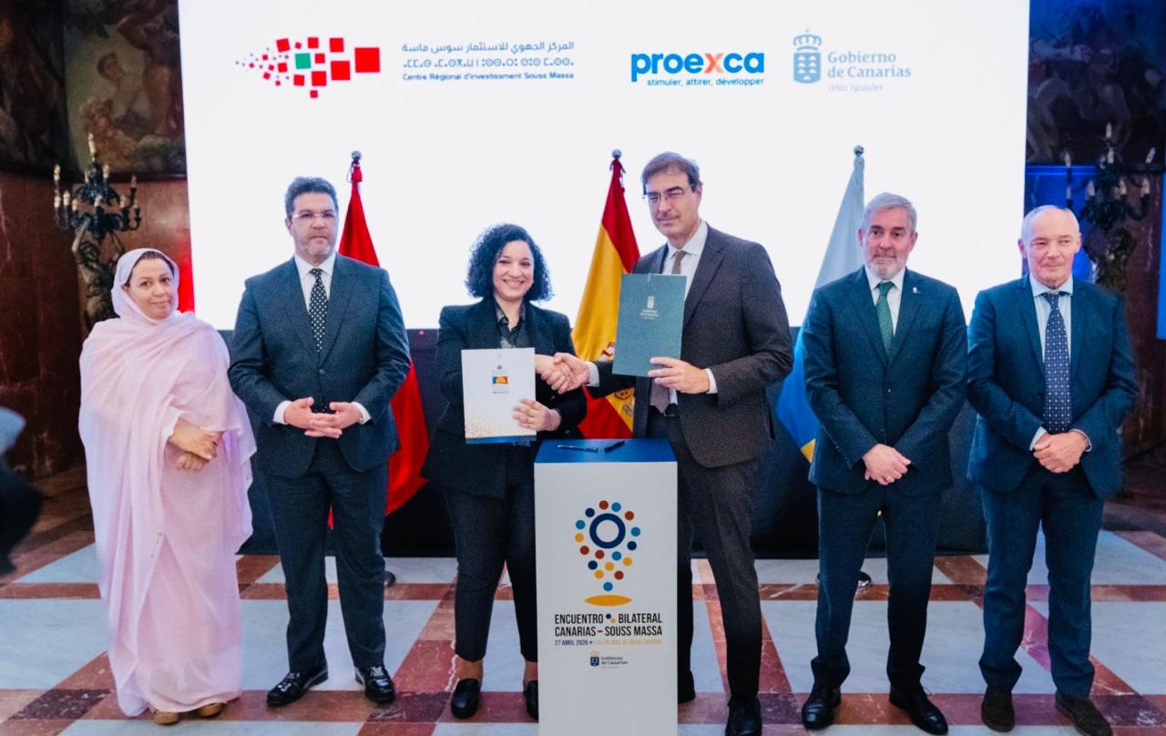 Six officials in suits stand during a signing ceremony; the center pair shake hands and hold documents, with flags and a banner behind them.