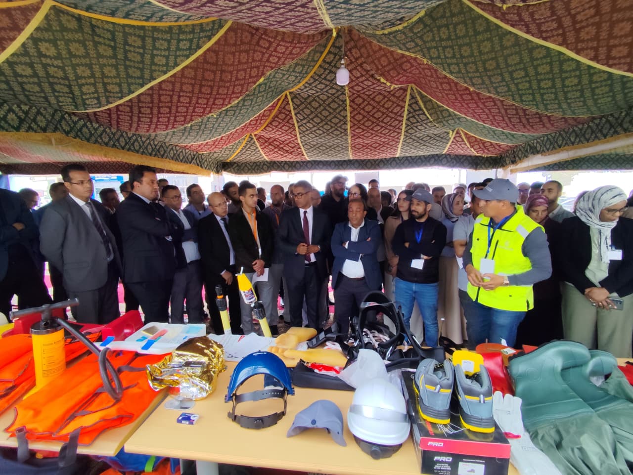 Group of professionals under a decorative canopy watching a display of safety gear on tables (helmets, life jackets, vests).