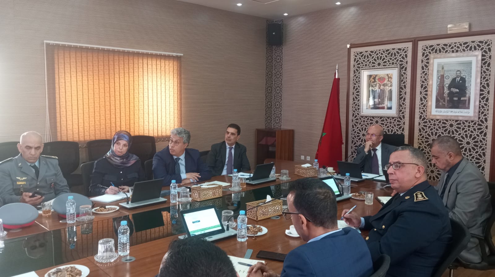 Group of officials in a conference room around a long table with laptops and bottled water; Moroccan flag in the background.