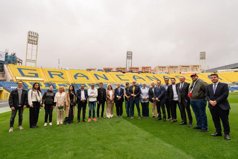 Group of people in business attire posing together on a soccer field inside a stadium, with yellow seats forming a background pattern behind them.