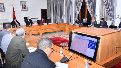 Formal committee meeting in a conference room with officials seated around a U-shaped wooden table; a large monitor on the foreground displays Arabic charts and notes while participants take notes.