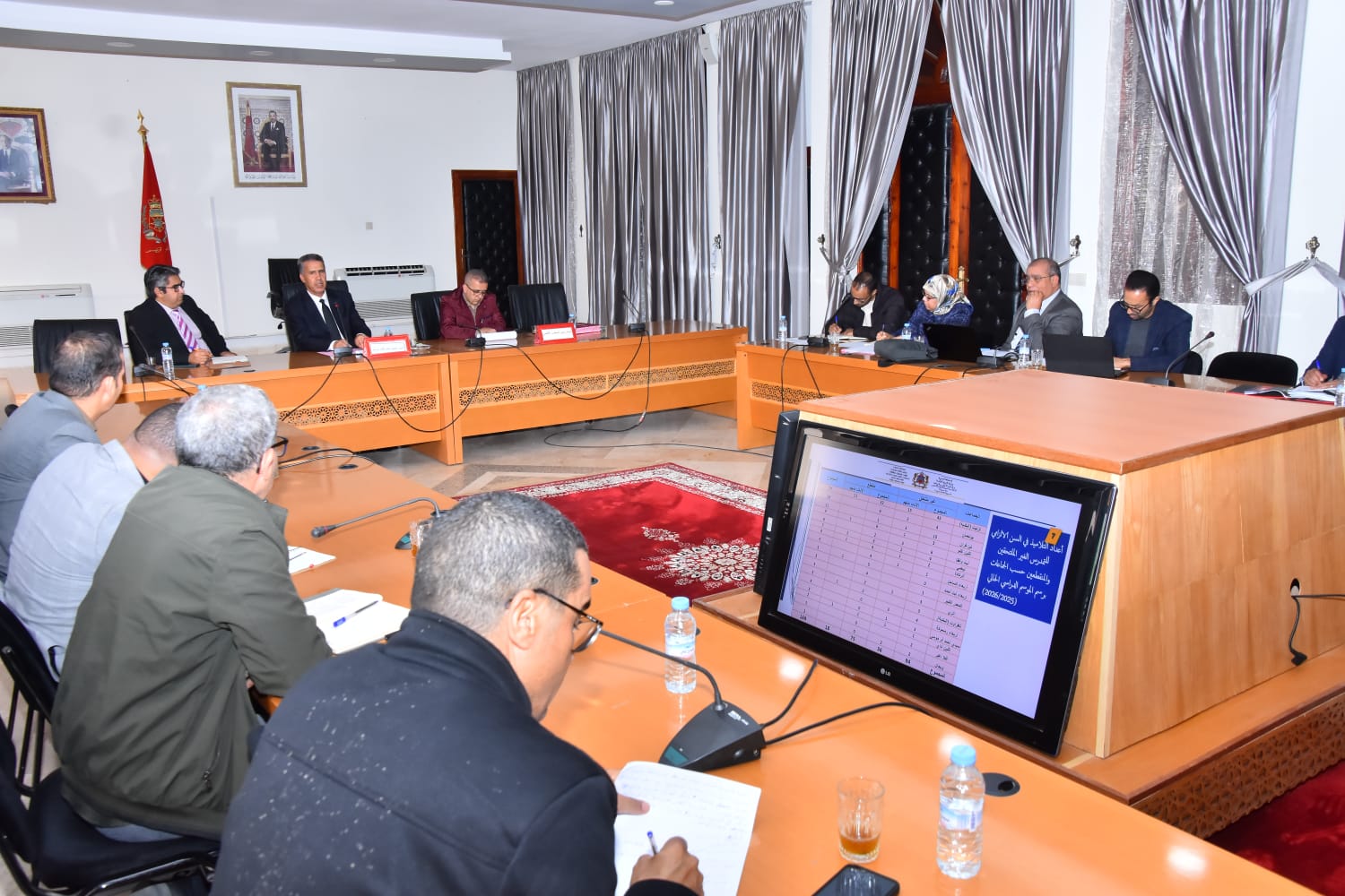 Formal committee meeting in a conference room with officials seated around a U-shaped wooden table; a large monitor on the foreground displays Arabic charts and notes while participants take notes.