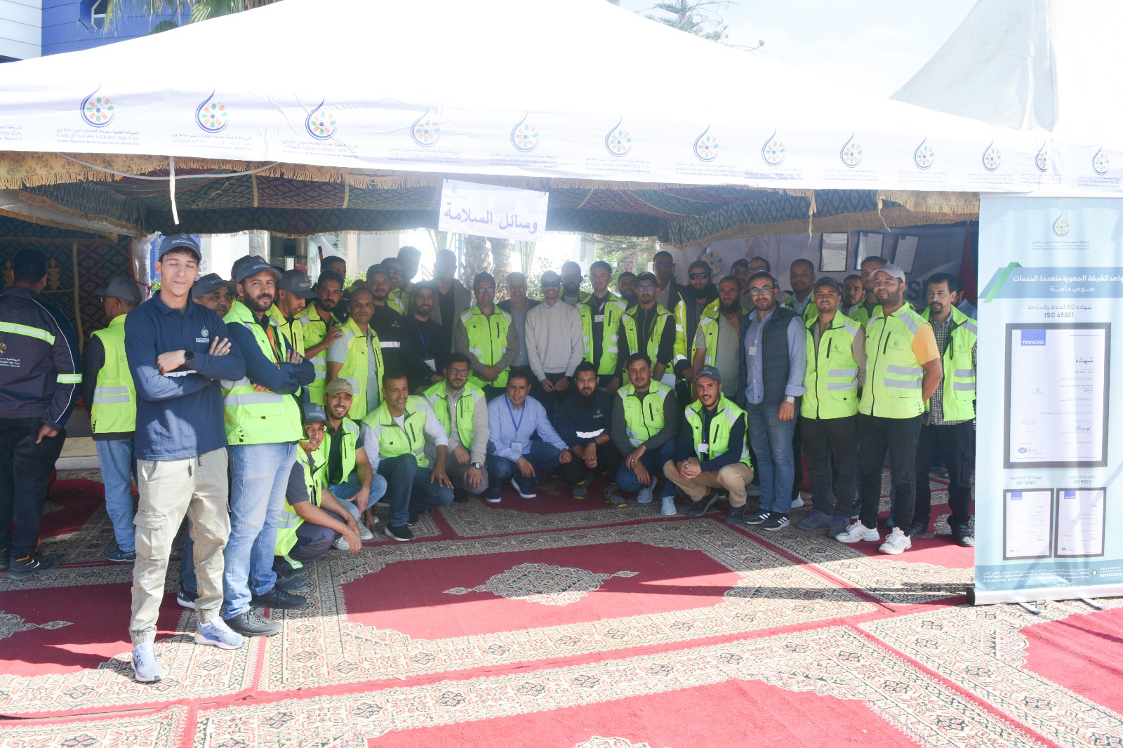 Group of workers wearing neon safety vests posing under a white canopy at an outdoor event.