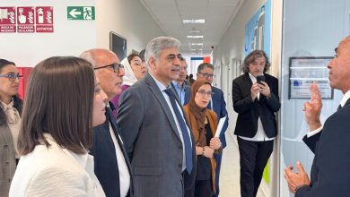 Group of professionals in a hallway listening to a speaker on the right; a man in a suit gestures as others, including a woman wearing a headscarf, observe.