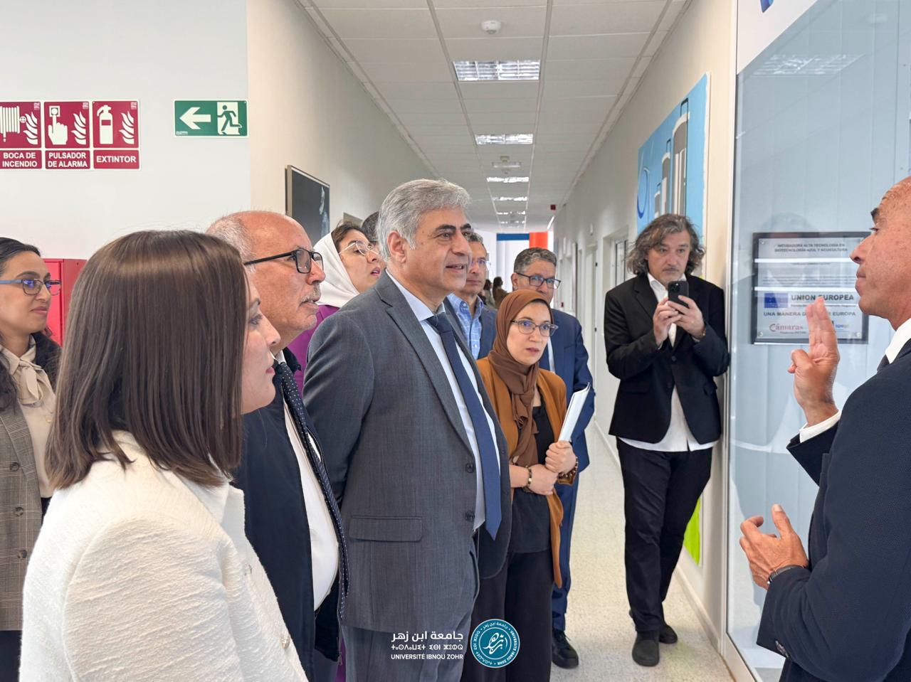 Group of professionals in a hallway listening to a speaker on the right; a man in a suit gestures as others, including a woman wearing a headscarf, observe.