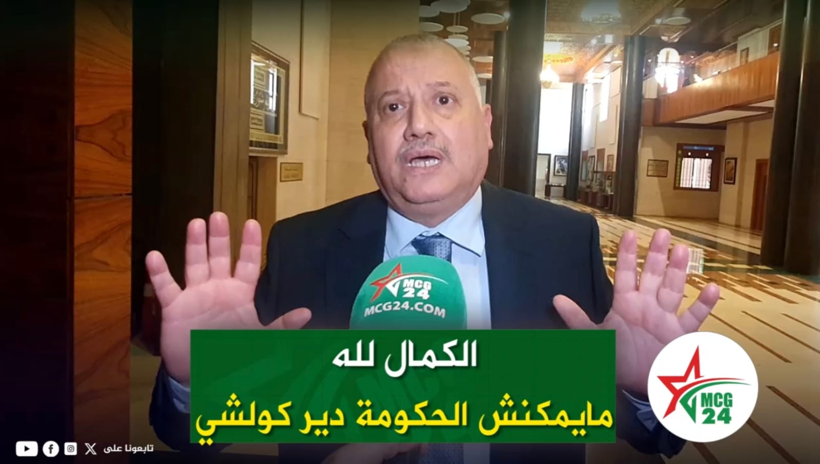 Man in a suit giving an interview in a marble lobby, hands raised, with a green Arabic caption and MCG24 microphone in front of him.