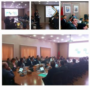 Professionals seated around a long conference table as a presenter speaks at the front of a meeting room with a screen displayed behind them