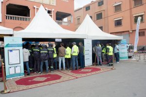 Group of people in reflective safety vests inside a white tent at an outdoor exhibition, with banners and signs visible nearby in a city square.