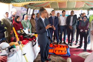 Man in a suit speaks to a group at a safety gear exhibit; PPE and rescue bags are on display.
