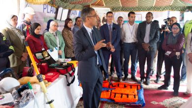 Man in a suit speaks to a group at a safety gear exhibit; PPE and rescue bags are on display.
