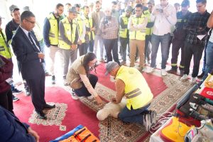 Group of people in hi-vis vests encircle a man kneeling to perform CPR on a dummy manikin during a training session on a red carpeted floor.