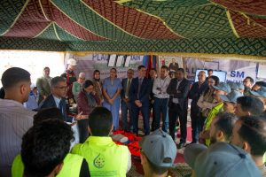 Group of officials and workers listening to a speaker at a training/ceremony under a decorative canopy, with life jackets laid out in front.