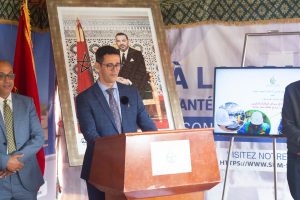 Man in a blue suit speaks at a wooden podium during a formal event, with a framed portrait and Moroccan flag behind him.