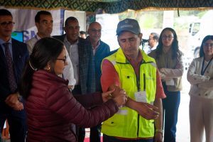 Woman in maroon jacket shakes hands with a man in a bright yellow safety vest at a group event indoors. Several onlookers watch nearby.