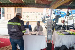 Man in a dark jacket with reflective stripes talks to two volunteers at an information booth under a colorful canopy, brochures on the table nearby.