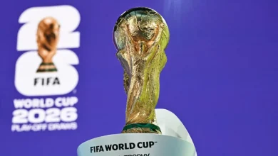 The FIFA World Cup trophy held upright with a blue backdrop and the 2026 event logo visible in the background