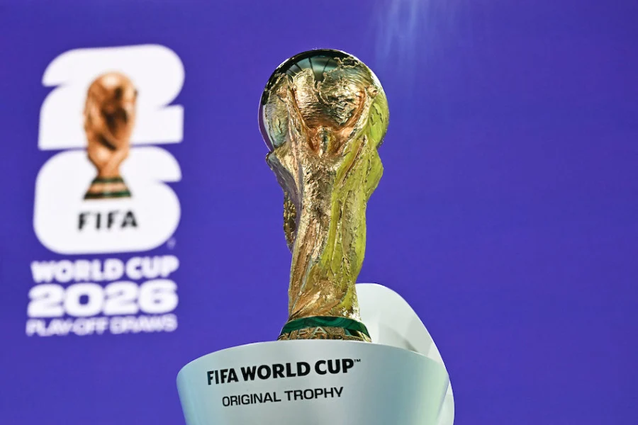 The FIFA World Cup trophy held upright with a blue backdrop and the 2026 event logo visible in the background