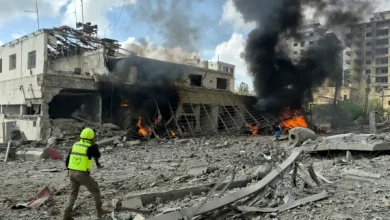 Rescue worker in a neon safety vest and helmet walks amid rubble as flames and black smoke rise from a destroyed building area.