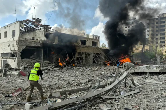 Rescue worker in a neon safety vest and helmet walks amid rubble as flames and black smoke rise from a destroyed building area.