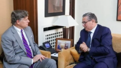 Two businessmen in suits sit in a wood-toned office, facing each other and talking.