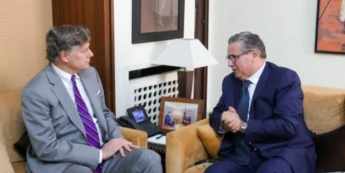 Two businessmen in suits sit in a wood-toned office, facing each other and talking.