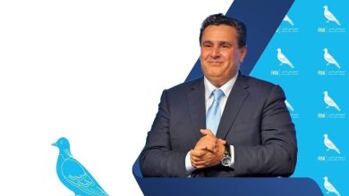 Man in a suit smiles while standing behind a blue podium at a press event with blue bird logos in the background.