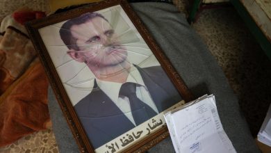 Framed portrait of a man in a suit with cracked glass, Arabic text at bottom, lying on a blanket with papers nearby.