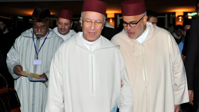 Three men in white robes and red fezzes walk together at an indoor event; the man in the center smiles while holding a booklet.