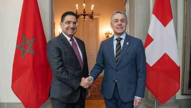 Two men in suits shake hands in a formal hallway, standing between the Moroccan flag (green star on red) and the Swiss flag (red with a white cross).