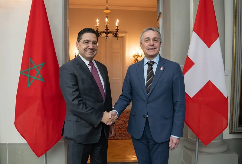 Two men in suits shake hands in a formal hallway, standing between the Moroccan flag (green star on red) and the Swiss flag (red with a white cross).