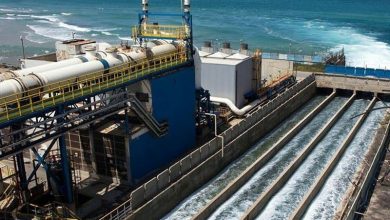 Industrial water treatment plant beside the ocean with large concrete channels carrying fast-flowing water, blue metal framework visible.