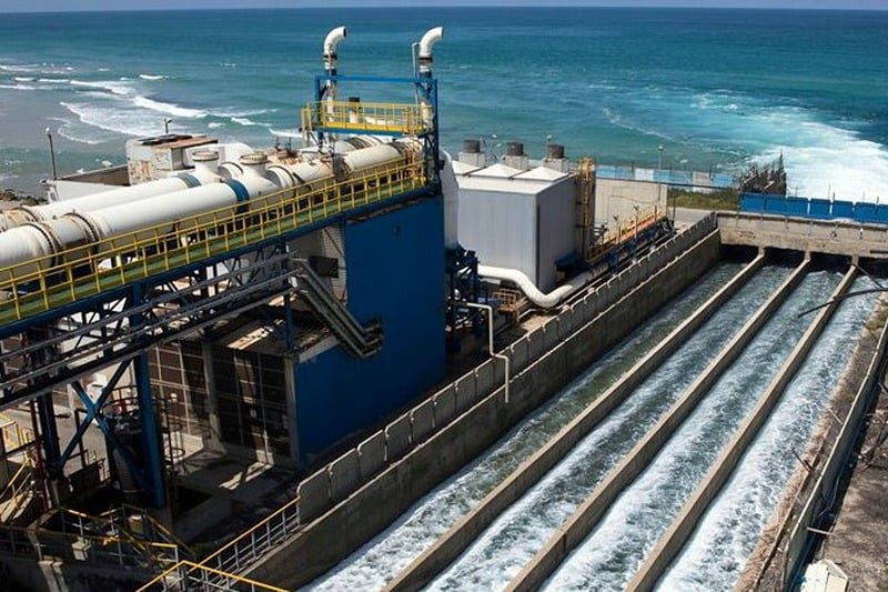 Industrial water treatment plant beside the ocean with large concrete channels carrying fast-flowing water, blue metal framework visible.