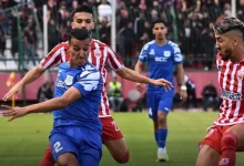 Soccer players in blue and red striped kits compete for the ball on a green field during a match at midfield moment.
