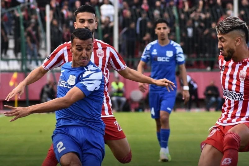 Soccer players in blue and red striped kits compete for the ball on a green field during a match at midfield moment.