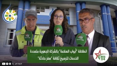 Three reporters with green MCG24 microphones stand outside a blue building, with an Arabic caption banner across the bottom.