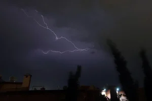 Dark stormy night with a jagged lightning bolt across the cloudy sky over city rooftops and tall trees in the foreground.