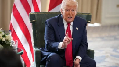 Man in a dark suit and red tie sits in a green chair, gesturing with one finger raised in a formal setting with flags in the background