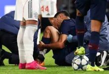 Soccer players surround an injured teammate on the field, one sitting with hands on his knee while others assist him.