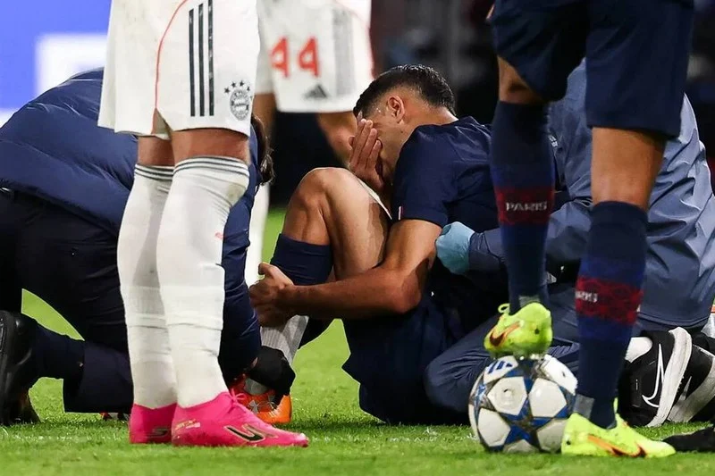 Soccer players surround an injured teammate on the field, one sitting with hands on his knee while others assist him.