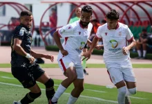 Three soccer players contest the ball during a match; two in white kits with Afriquia logos and red trim face a black-clad opponent.