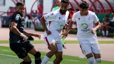 Three soccer players contest the ball during a match; two in white kits with Afriquia logos and red trim face a black-clad opponent.