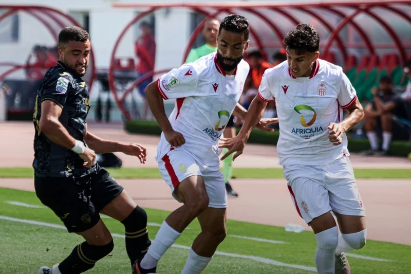 Three soccer players contest the ball during a match; two in white kits with Afriquia logos and red trim face a black-clad opponent.